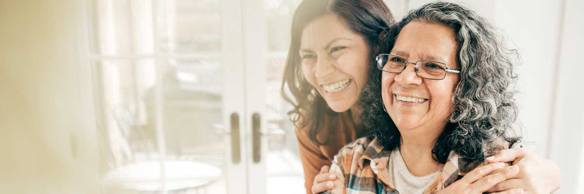 A young woman hugging an older woman from behind, both smiling